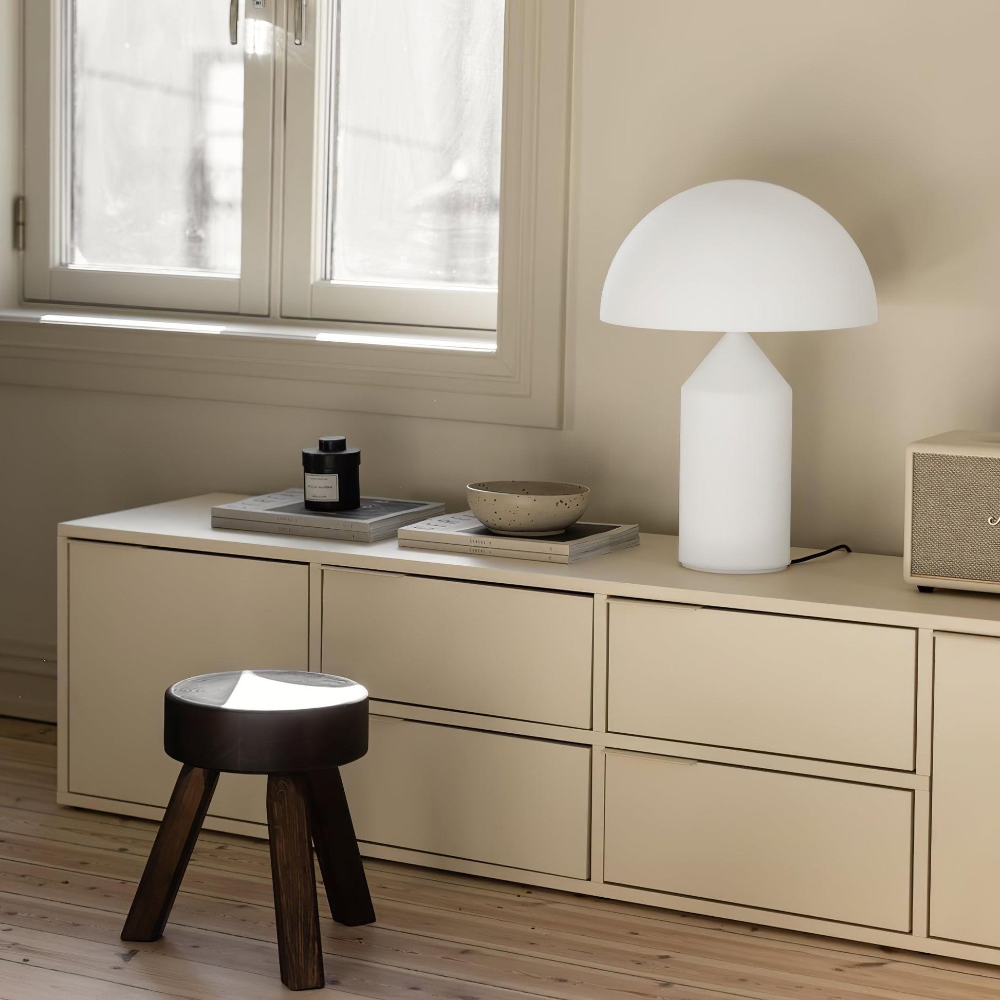 Modern interior with a white table lamp, wooden stool, and decorative items on a beige cabinet.