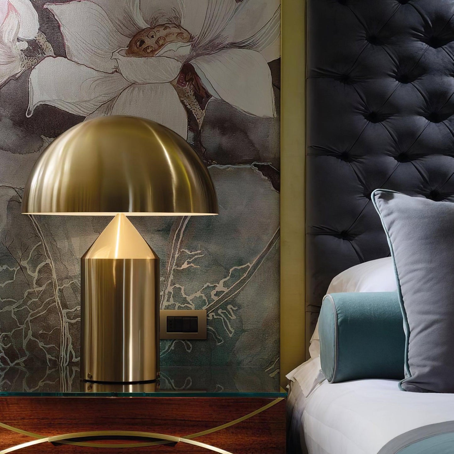 Gold table lamp on a nightstand next to a bed with floral wallpaper