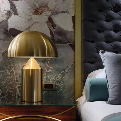 Gold table lamp on a nightstand next to a bed with floral wallpaper