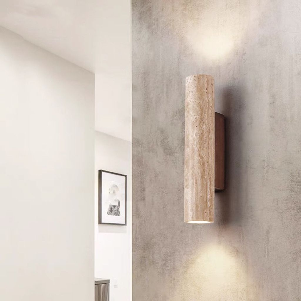 Wall-mounted cylindrical light fixture on a textured wall with a mirror and framed picture in the background.