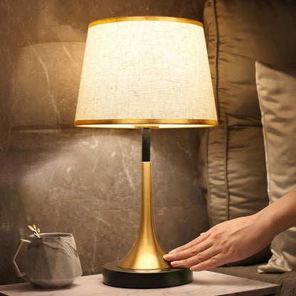 Table lamp with gold base and white shade on a table, hand reaching towards it.