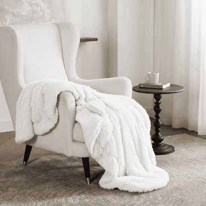 White plush blanket draped over a white armchair in a room with a side table and curtains.