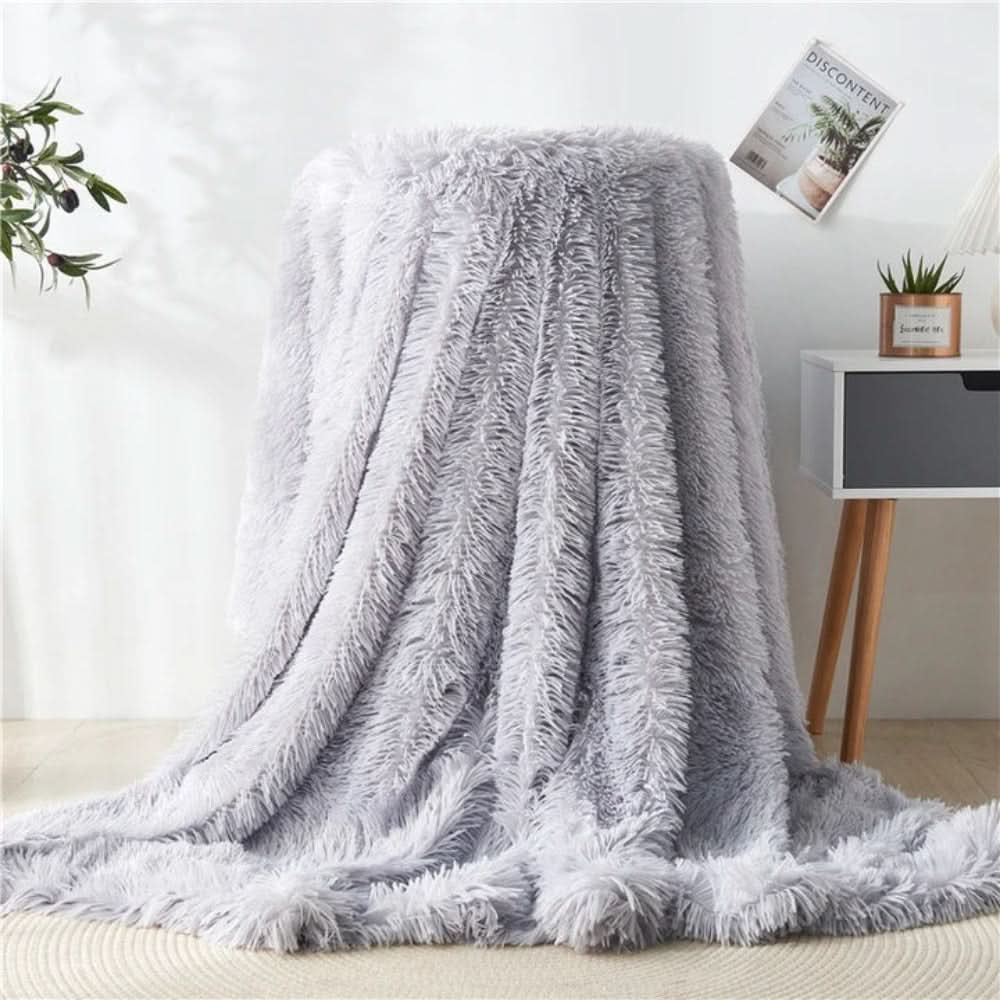 Fluffy gray blanket draped over a chair in a room with a plant and small table in the background.