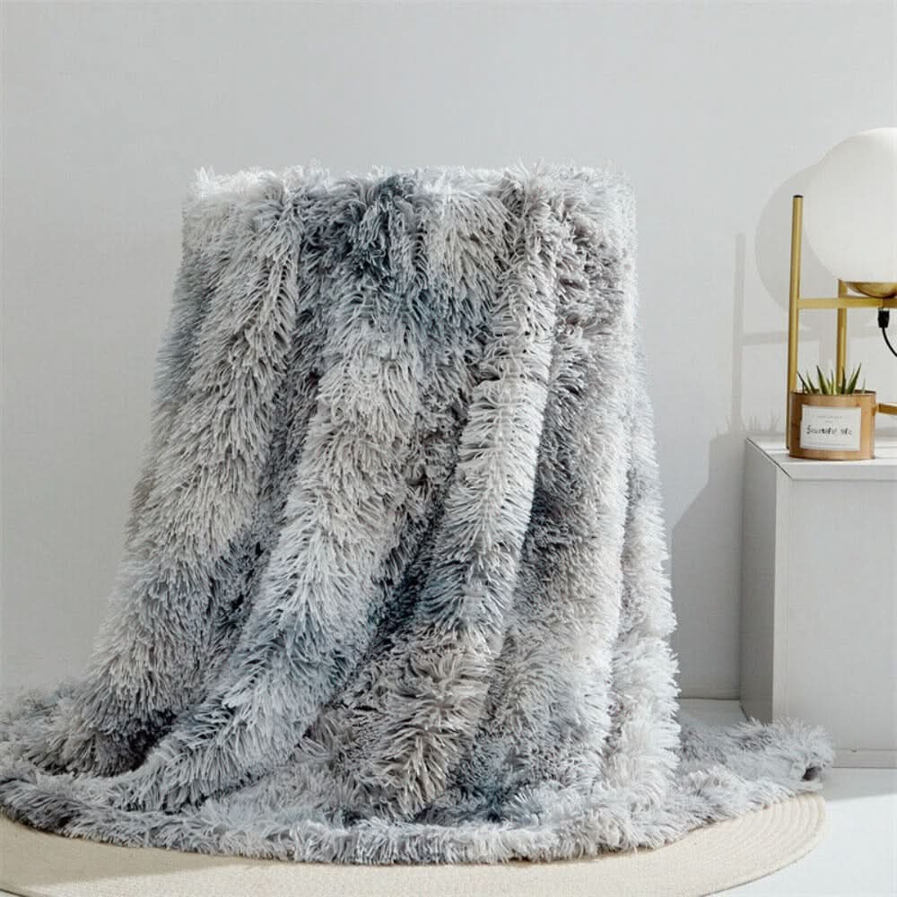 Fluffy gray blanket draped over a chair in a room with a lamp and plant in the background.