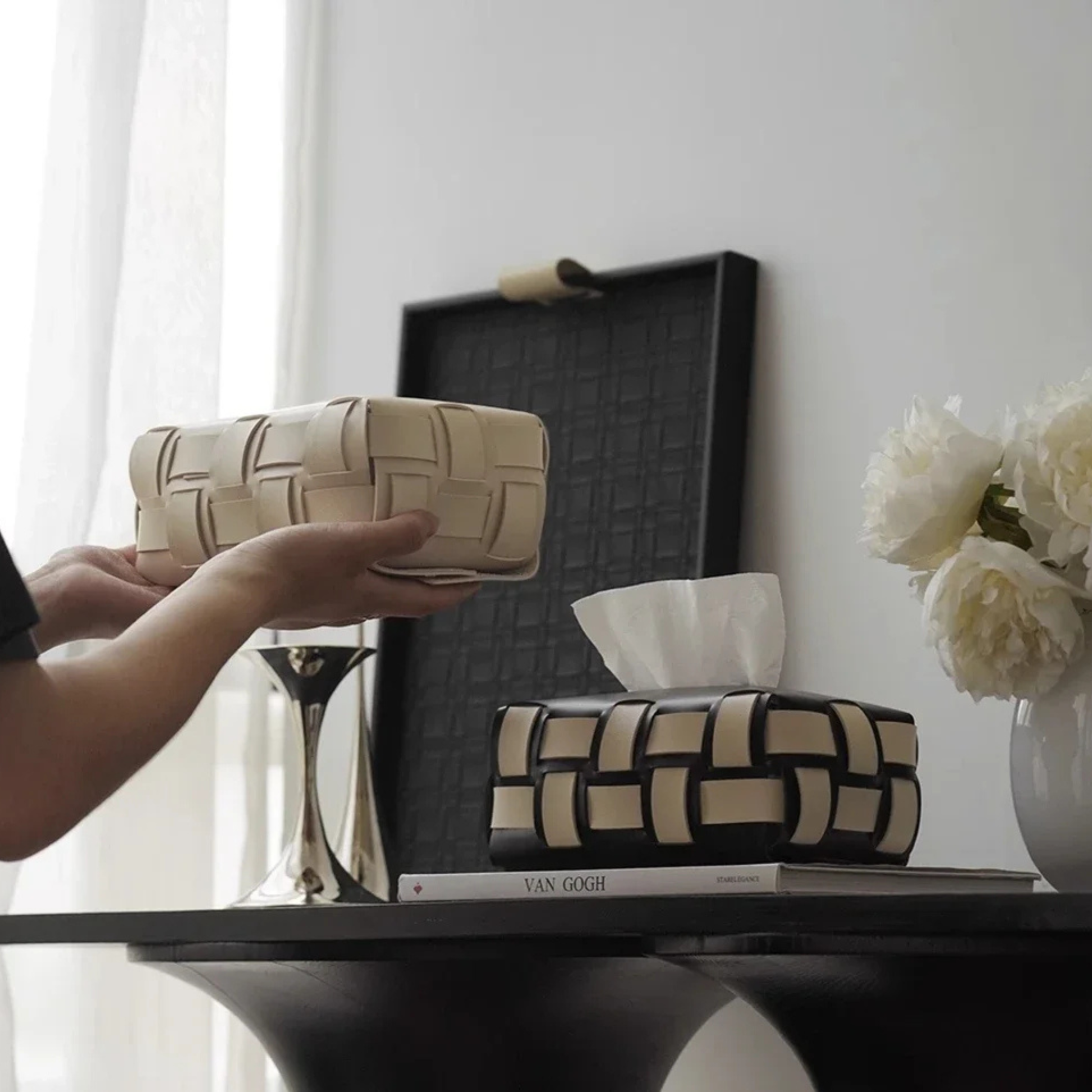 Person holding a woven tissue box cover on a table with decor items in the background