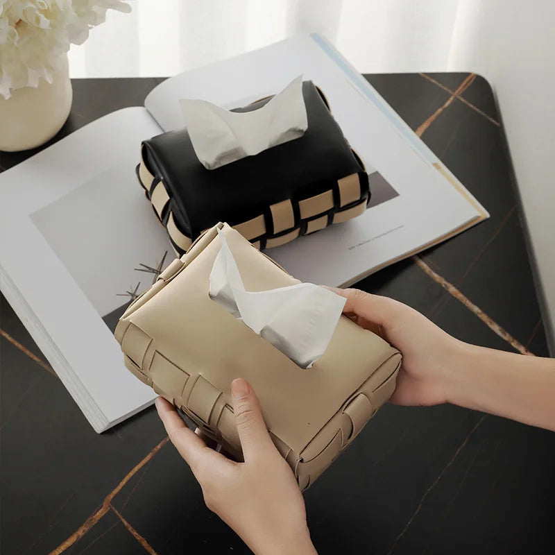 Beige tissue holder with tissues being held by a hand on a dark surface with a book and tissues in the background.