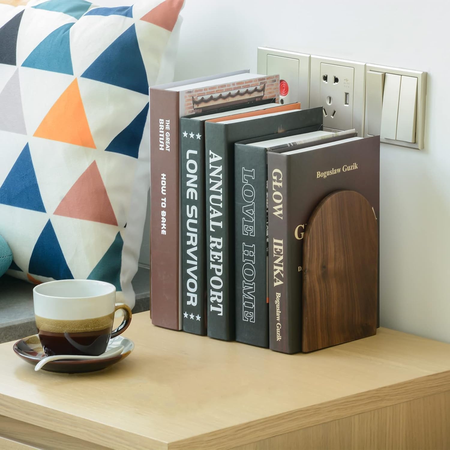 Stack of books on a wooden surface with a cup and saucer, next to a geometric-patterned cushion.