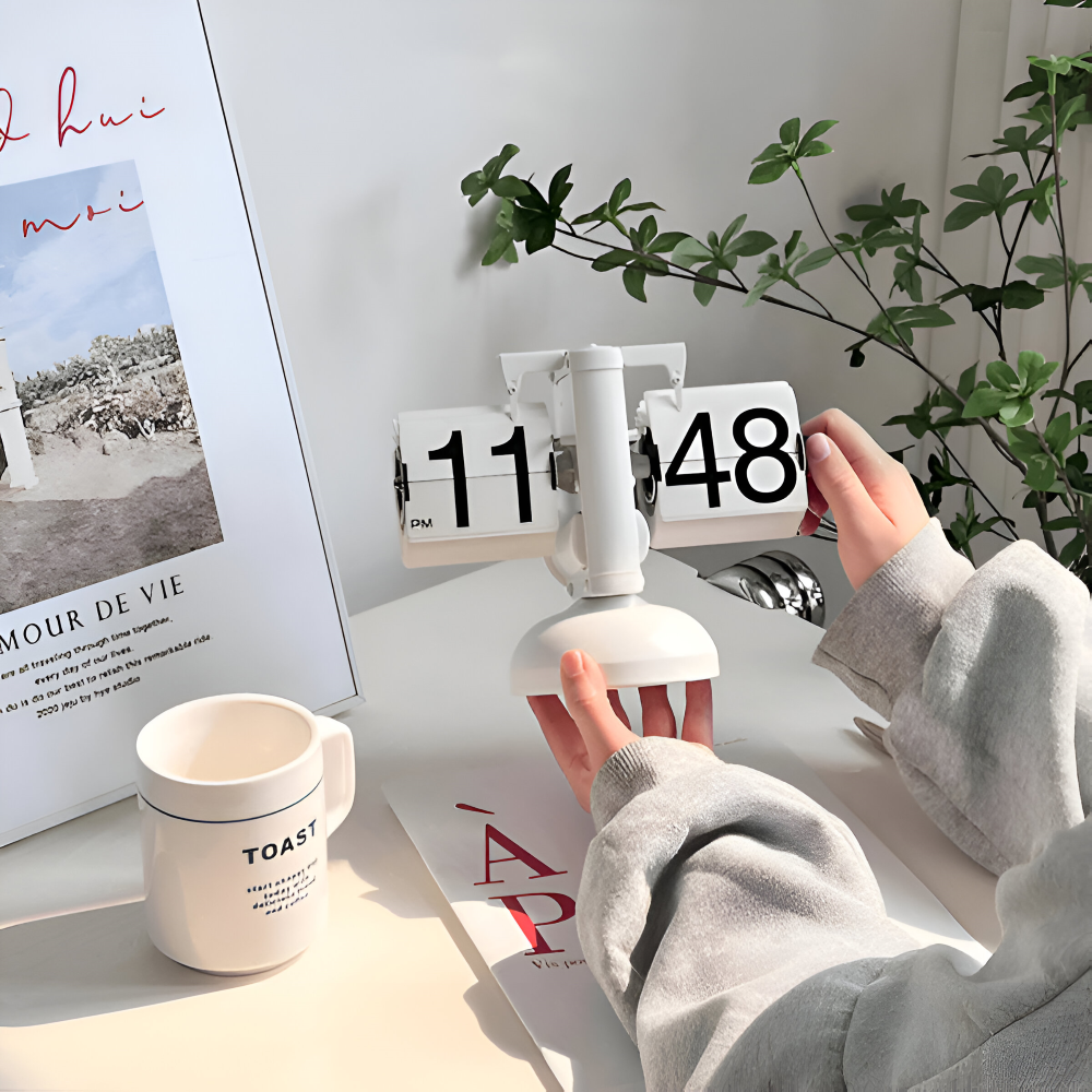 Person adjusting a white flip clock on a desk with a mug and plant in the background