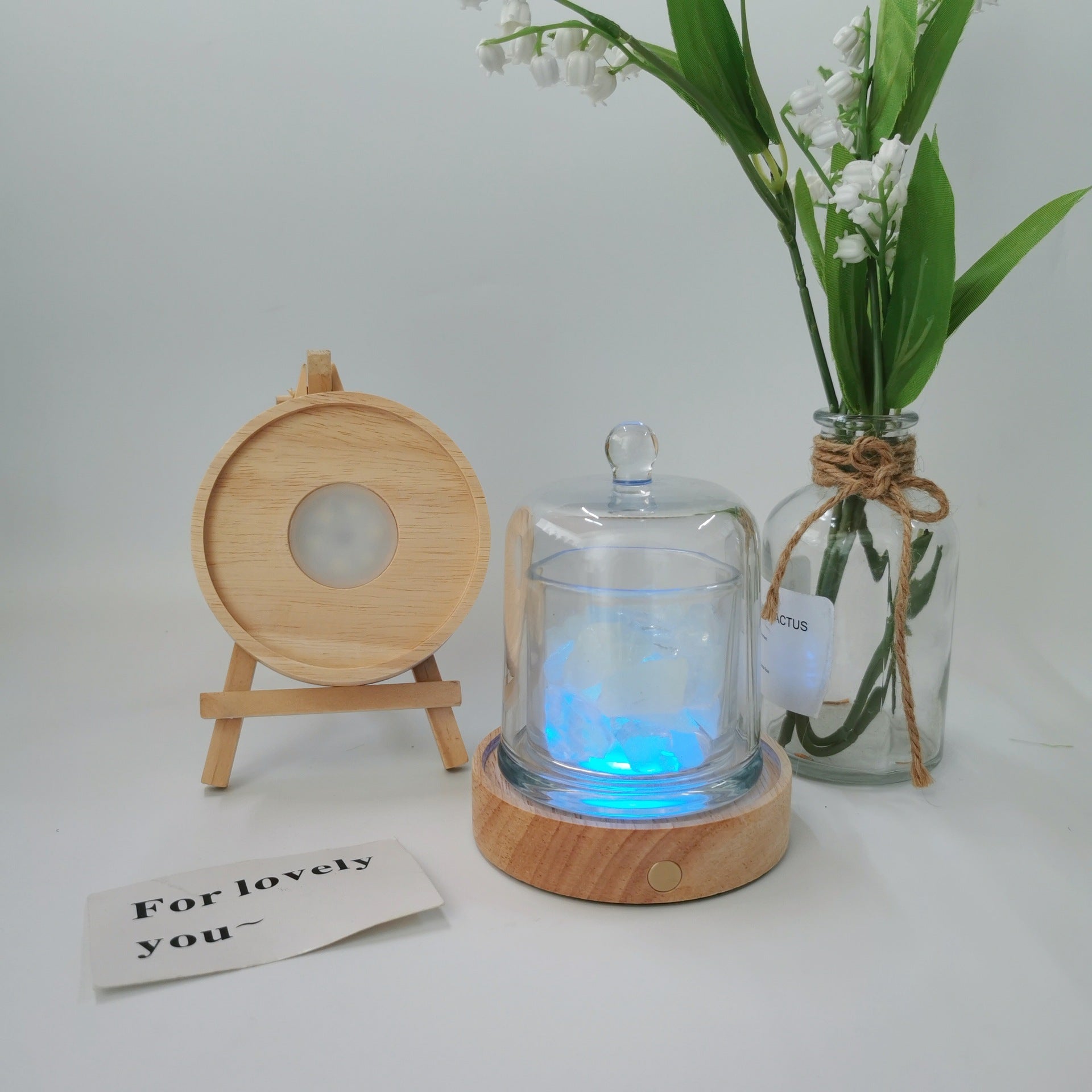 Glass diffuser with wooden base and light, next to a vase with flowers and a small card on a white background
