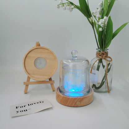 Glass diffuser with wooden base and light, next to a vase with flowers and a small card on a white background