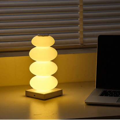 Stacked cylindrical lamp on a desk next to a laptop with blinds in the background