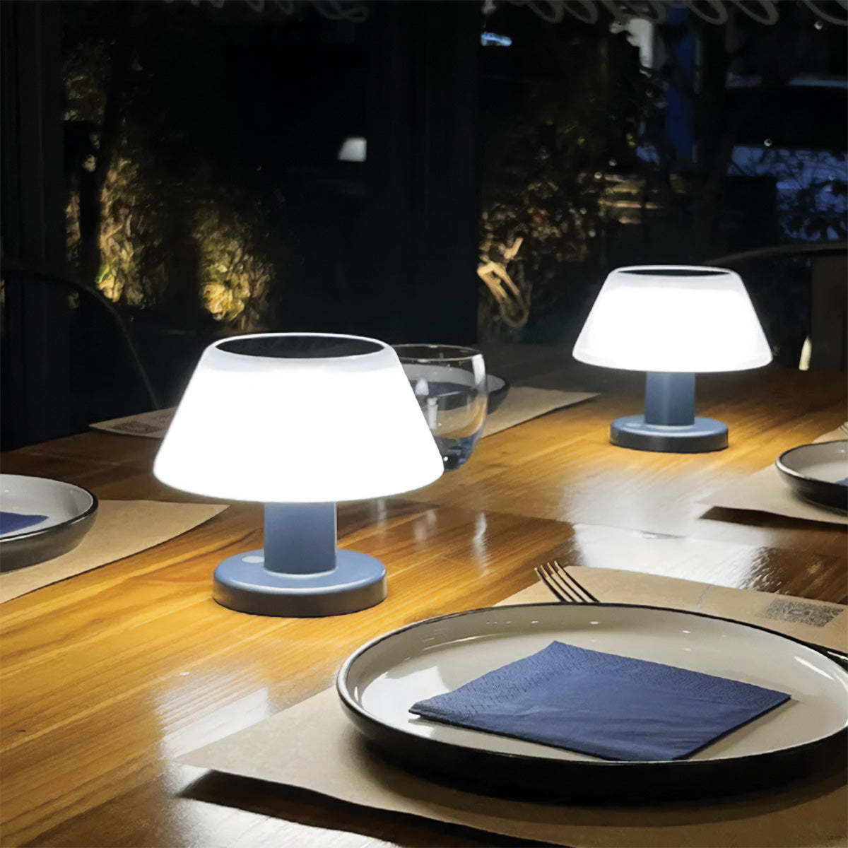Two small table lamps on a wooden table with plates and cutlery in the background.