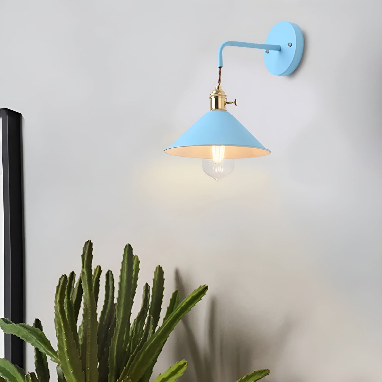 Wall-mounted light fixture with a blue shade and gold base on a white wall, next to a cactus plant.