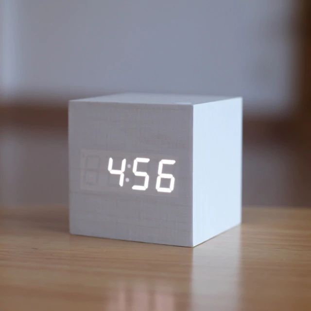 White digital clock displaying the time on a wooden surface with a blurred background